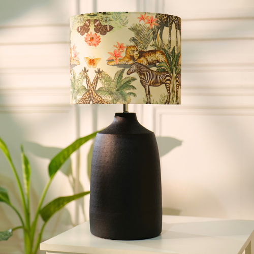 Table lamp with a dark, textured metal base and a floral-patterned fabric shade, placed on a white table in a room with plants